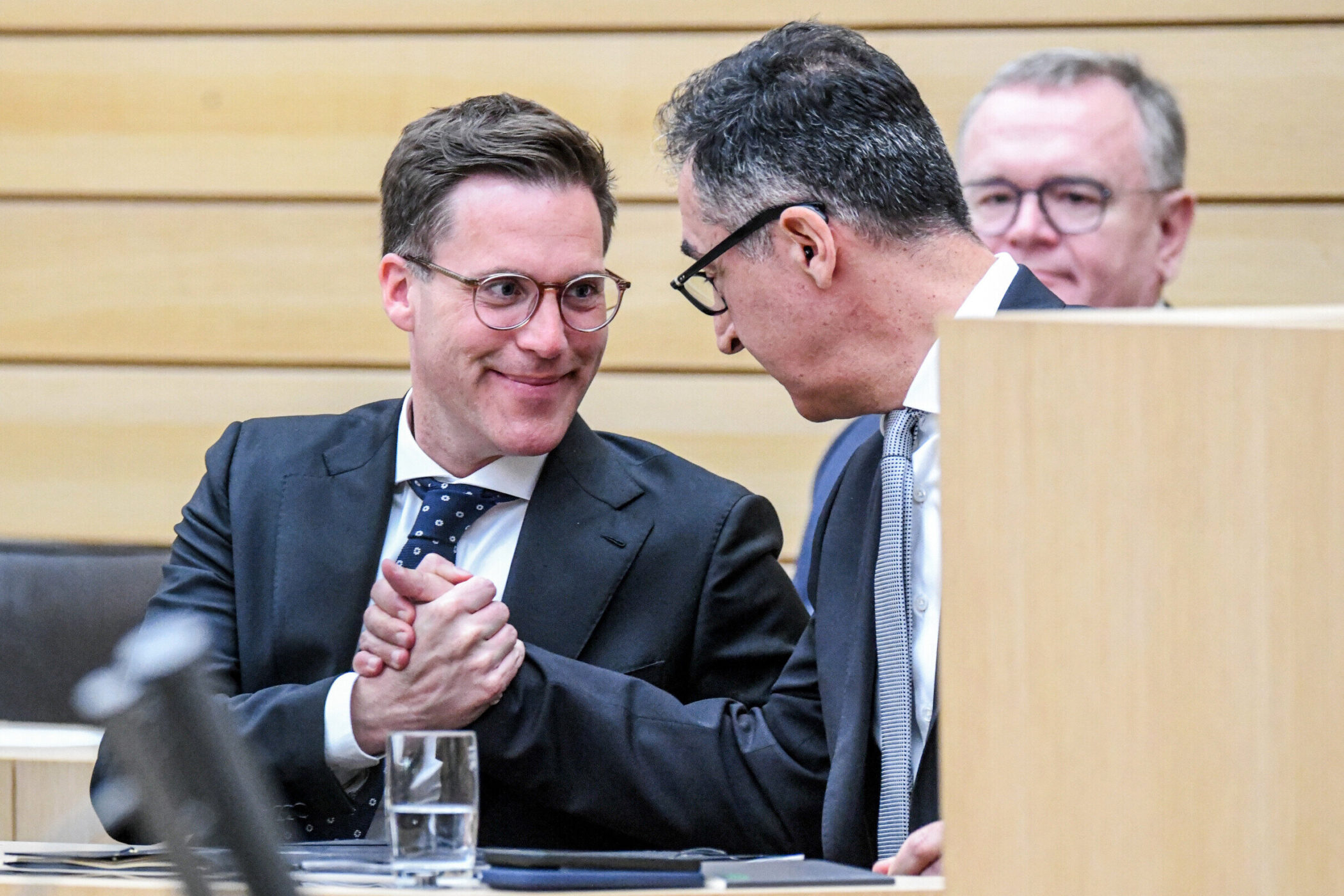 Drei Männer in Anzügen, zwei geben sich die Hand, einer im Hintergrund.