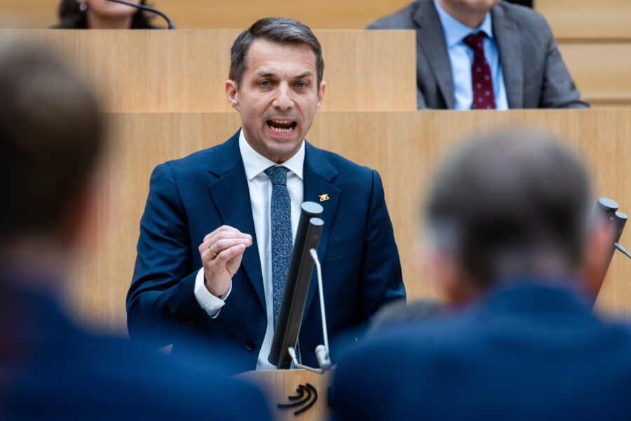 Mann in Anzug spricht am Rednerpult in einem Saal, weitere Personen im Hintergrund.