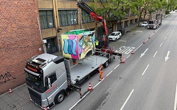 LKW mit Kran hebt bemalten Container, Bauarbeiter sichern die Straße ab.
