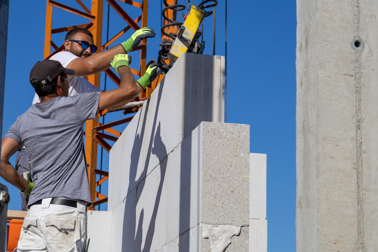 Zwei Männer bauen mit Zementblöcken und Krangerät eine Wand.