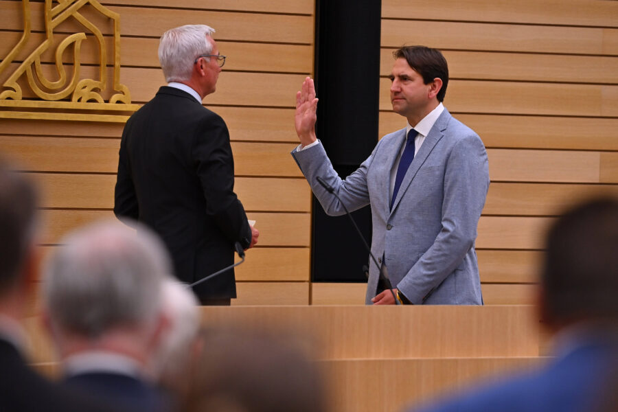 Zwei Männer in Anzügen stehen sich vor Holzwand gegenüber, einer hebt die Hand.