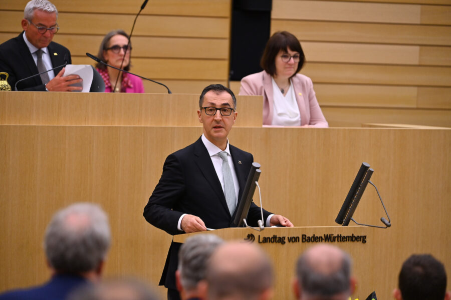 Mann spricht am Rednerpult im Landtag von Baden-Württemberg, drei Personen im Hintergrund.
