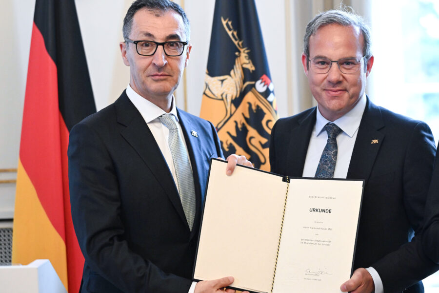 Zwei Männer halten eine Urkunde vor den Fahnen von Deutschland und Baden-Württemberg.