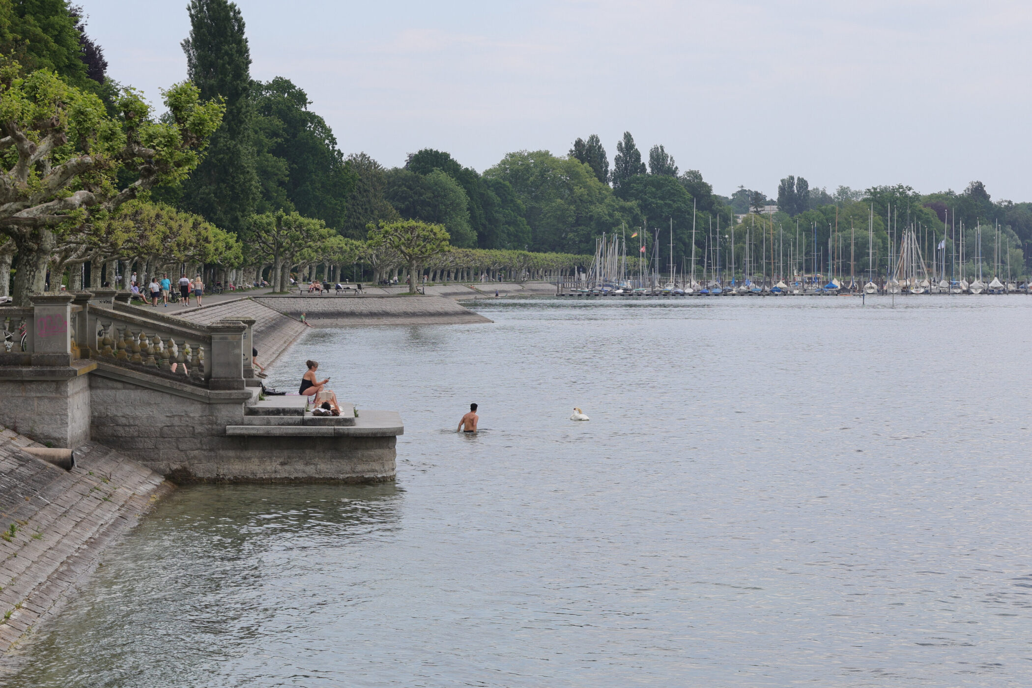 Uferpromenade mit Spaziergängern, badendem Mann und im Hintergrund viele Boote.