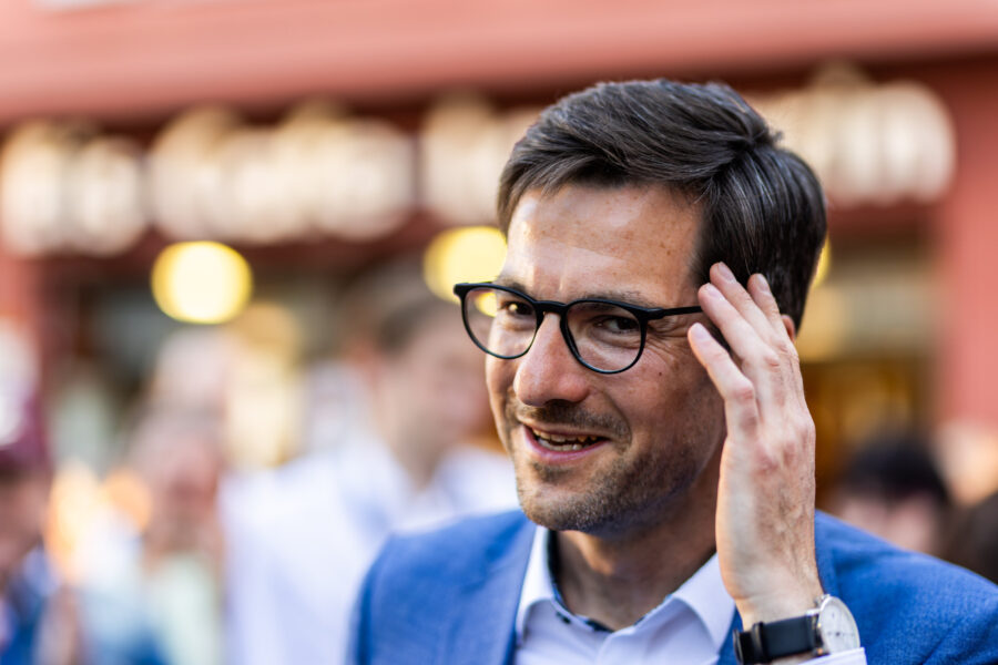 Mann mit Brille in blauem Anzug, berührt sein Haar, verschwommener Hintergrund.