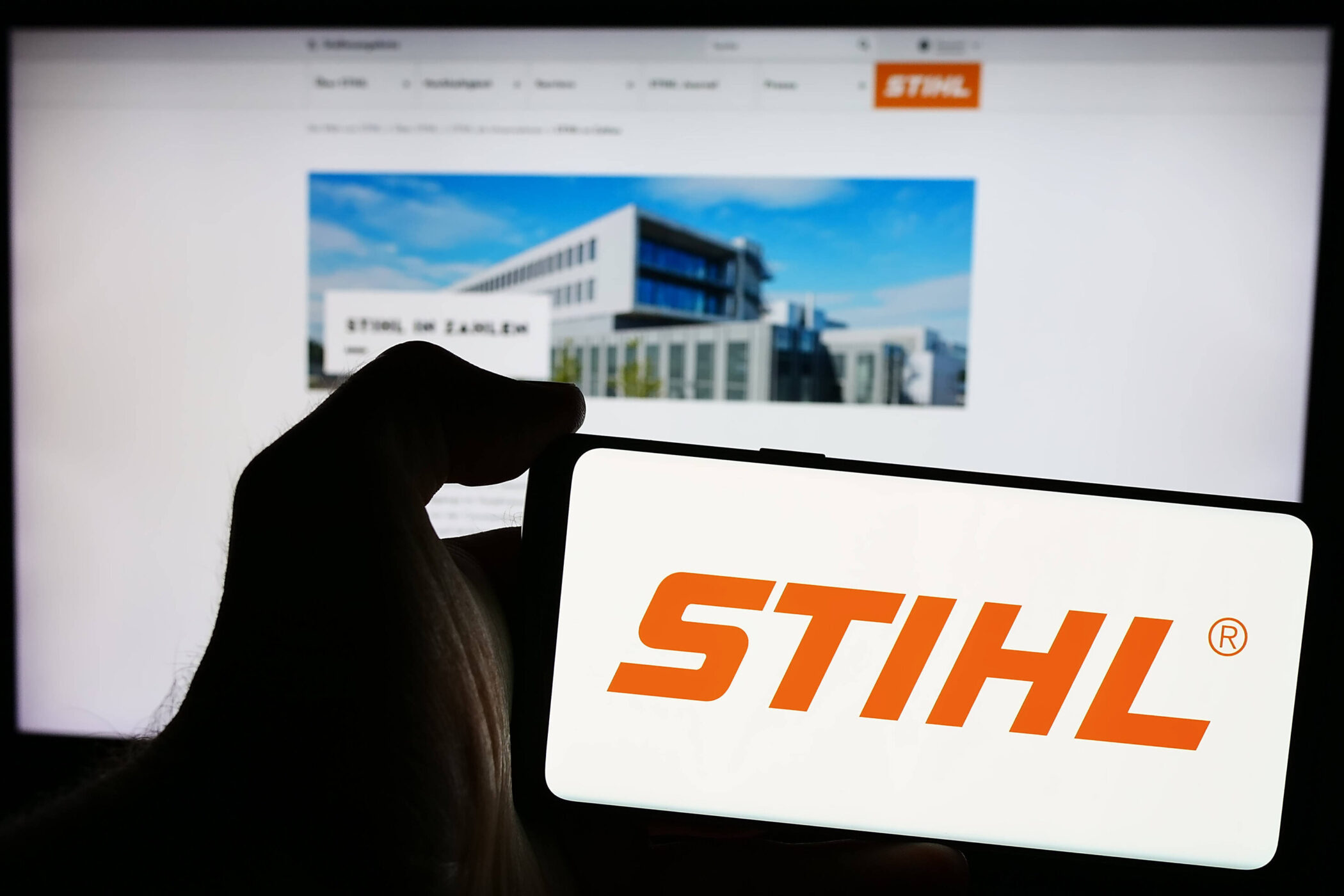 Hand hält Smartphone mit STIHL-Logo, im Hintergrund unscharfer Bildschirm.