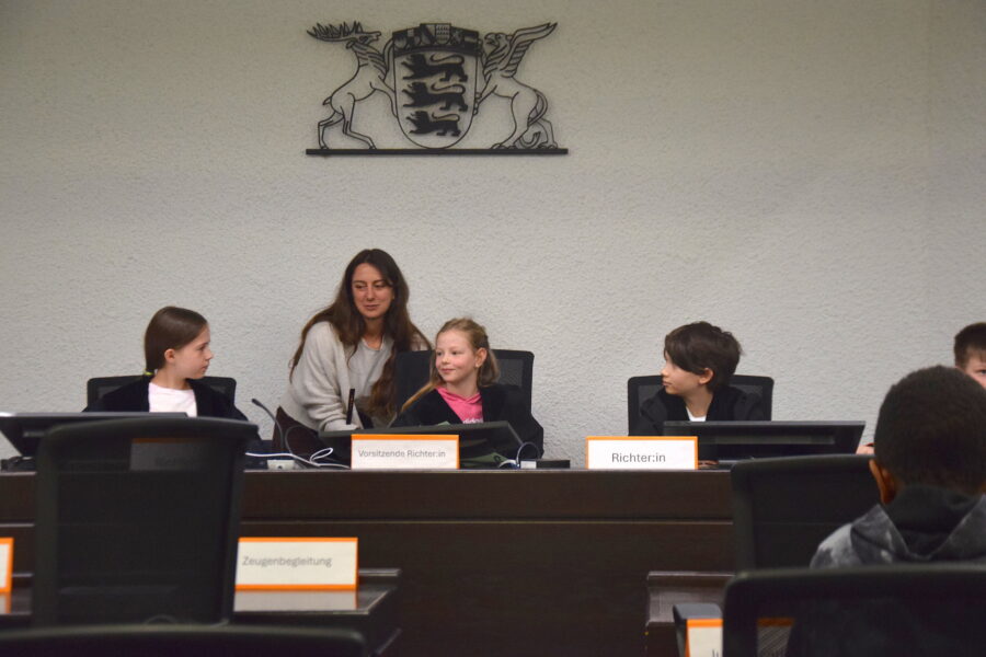 Fünf Personen in einem Raum mit Namensschildern vor sich. Wappen an der Wand.