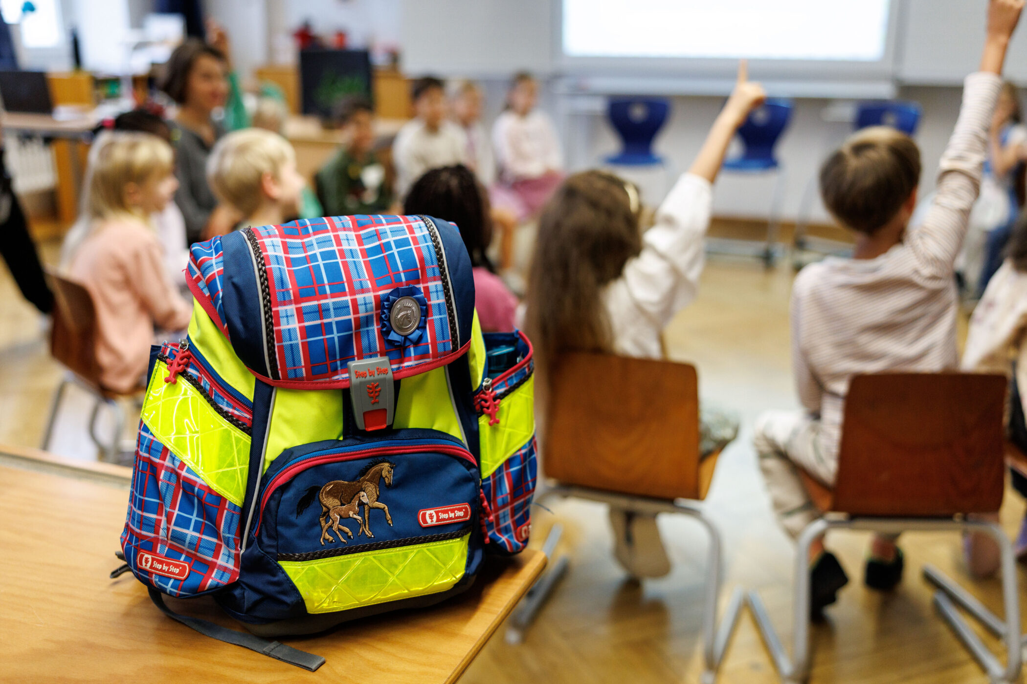 Schulrucksack im Vordergrund, Kinder auf Stühlen im Hintergrund.