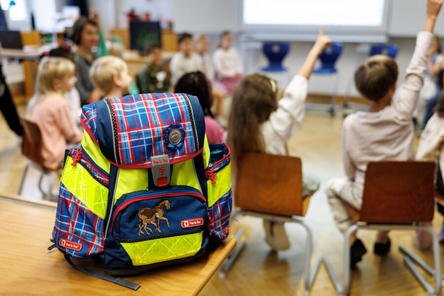 Schulrucksack im Vordergrund, Kinder auf Stühlen im Hintergrund.