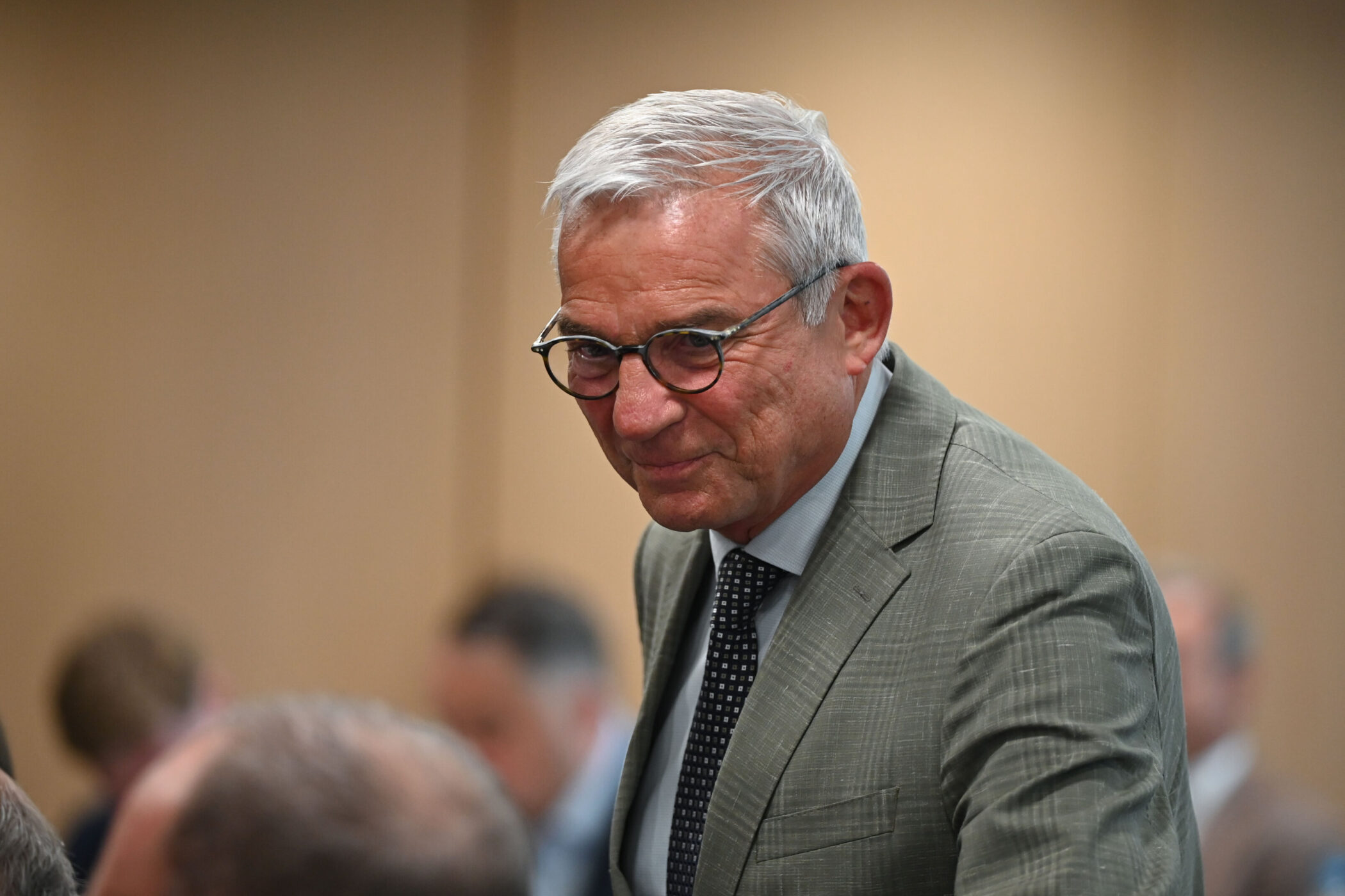 Älterer Mann mit Brille im Anzug vor unscharfem Hintergrund.