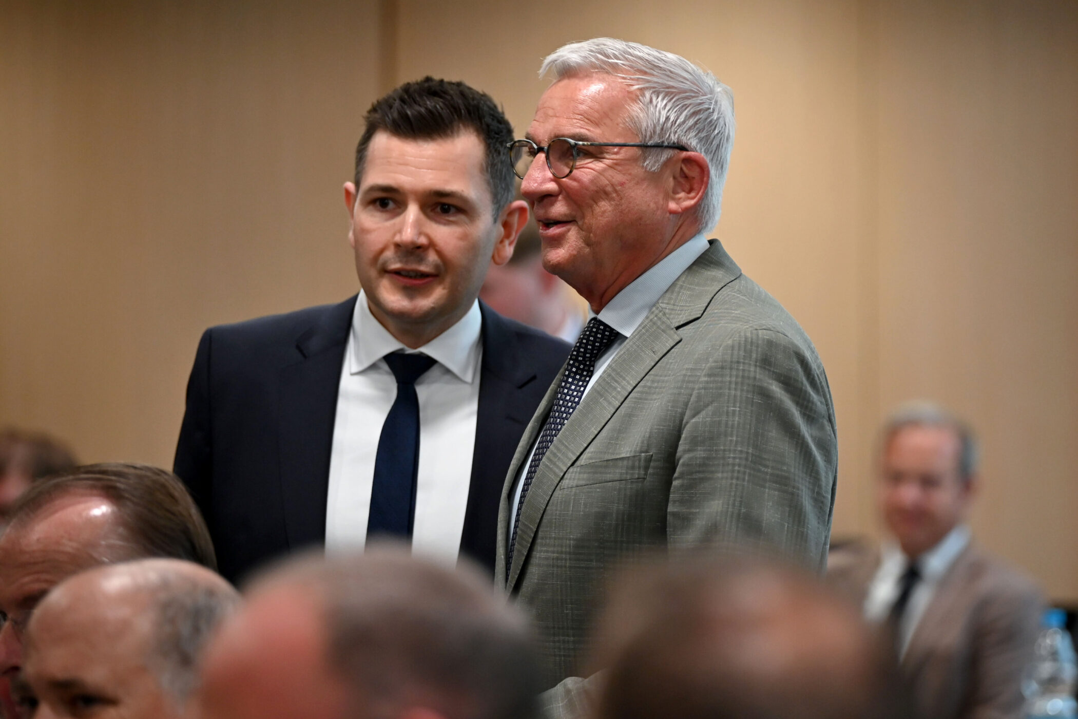 Zwei Männer in Anzügen im Gespräch, unscharfe Zuhörer im Hintergrund.