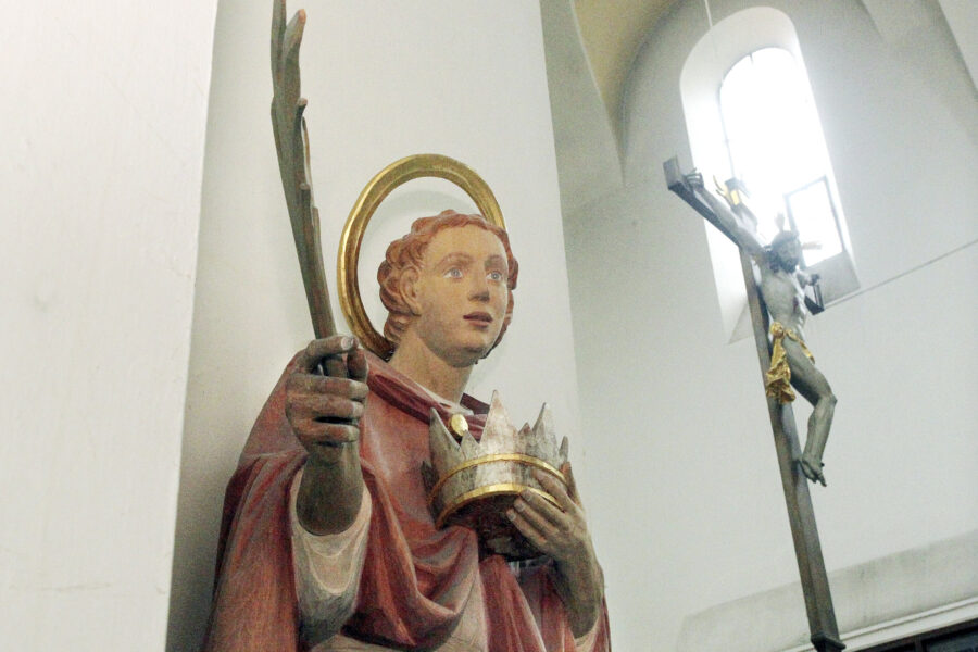 Statue mit Heiligenschein und Krone, im Hintergrund Kruzifix an der Wand.