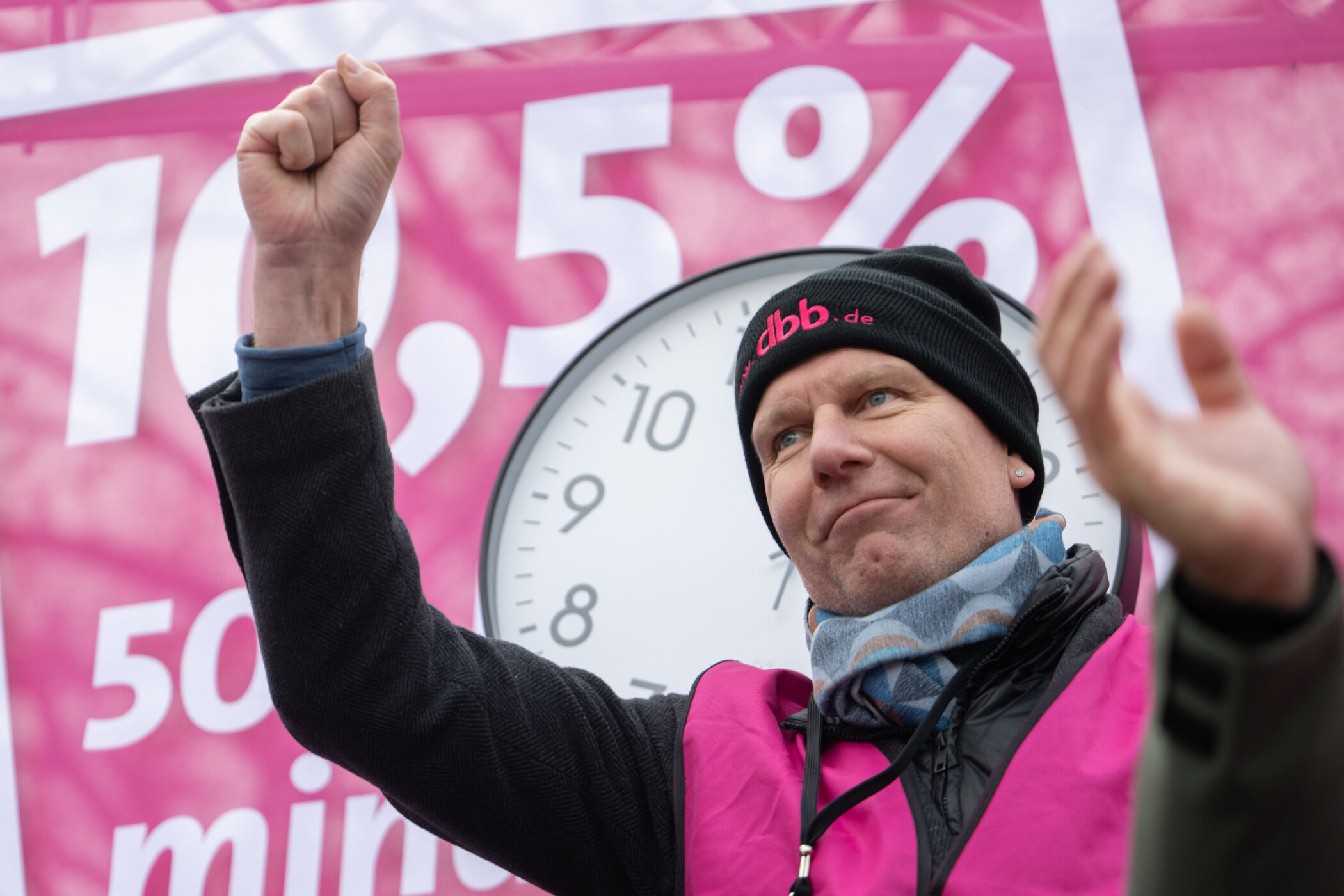 Mann mit erhobener Faust, pinker Hintergrund mit 10,5 %, große Uhr im Hintergrund.