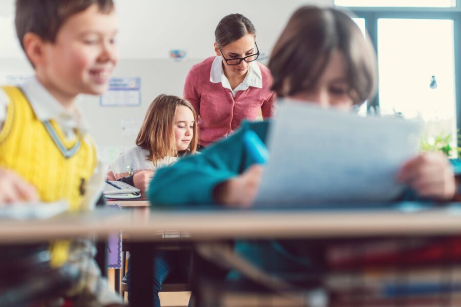 Mehrere Kinder sitzen an Tischen, eine Frau steht daneben, alle im Klassenraum.