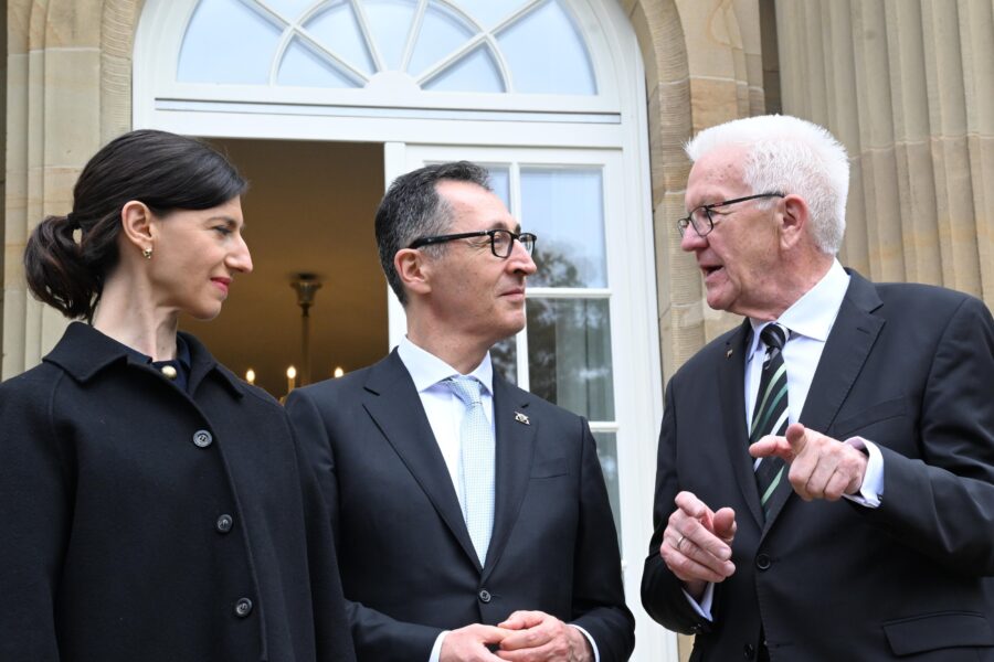 Drei Personen in Anzügen stehen vor einem Gebäude und unterhalten sich.