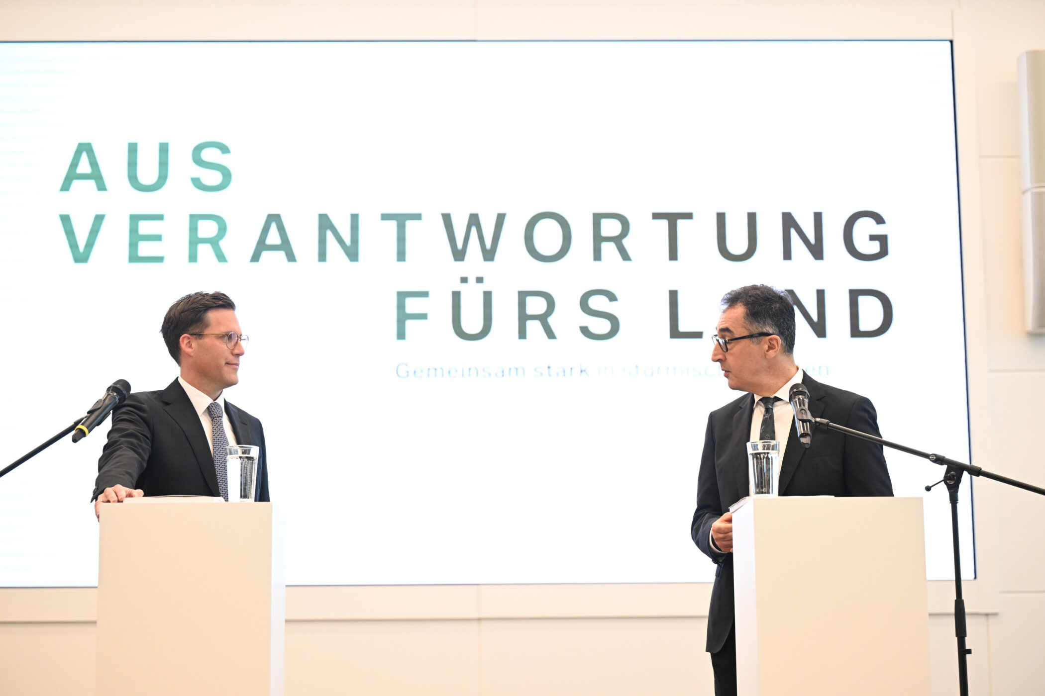 Zwei Männer in Anzügen stehen an Rednerpulten vor einem Bildschirm mit Text.