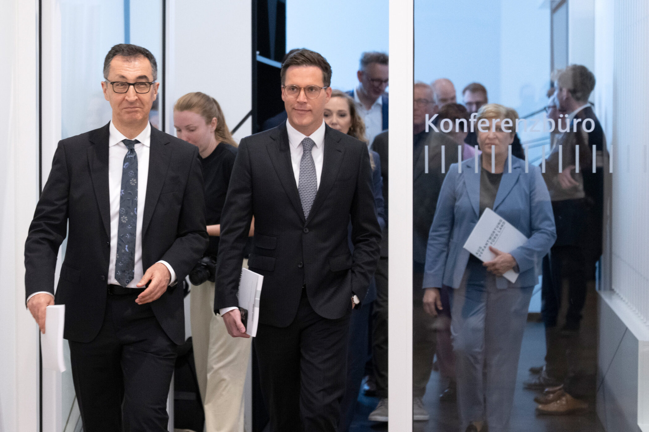 Mehrere Personen in Anzügen gehen einen Flur entlang. Türen sind mit "Konferenzbüro" beschriftet.