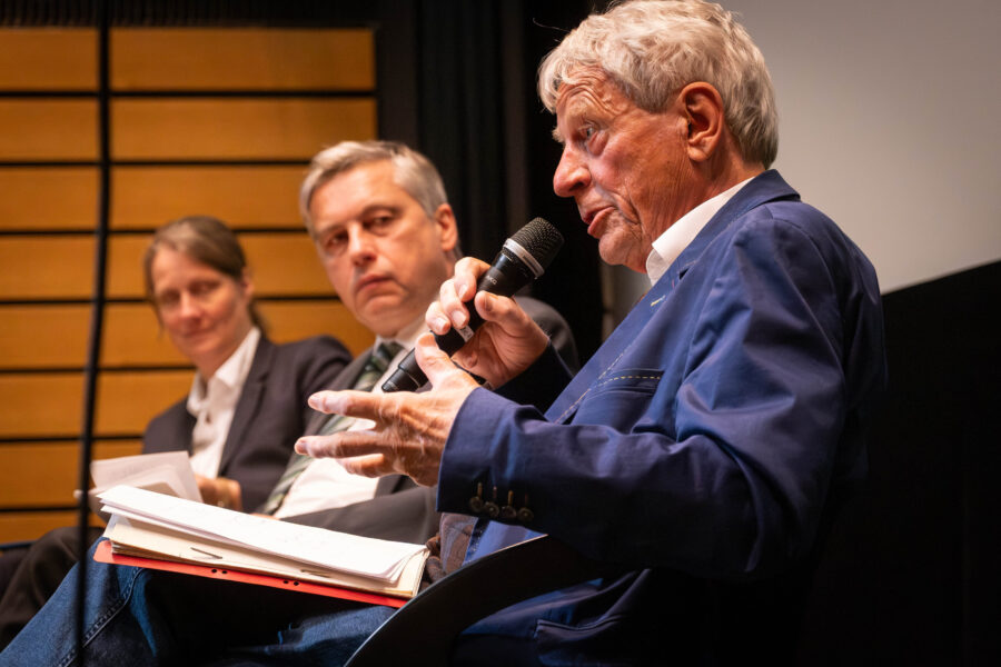 Drei Personen sitzen, eine Person spricht ins Mikrofon, Tisch mit Notizen im Vordergrund.