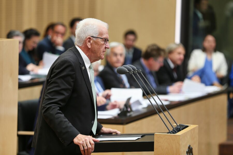 Ein Mann spricht an einem Rednerpult, Personen sitzen im Hintergrund.