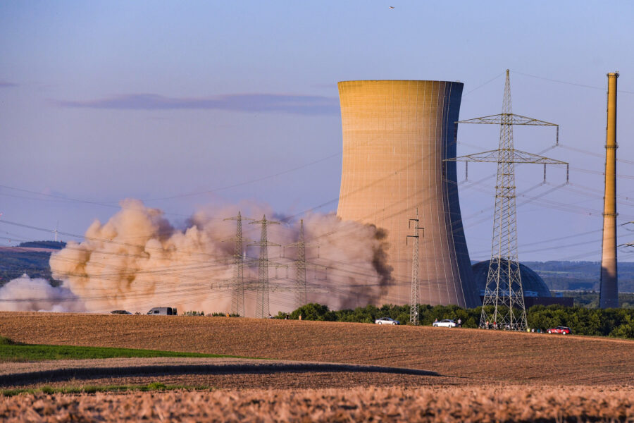 Rauchwolke steigt neben Kühlturm, Strommasten im Vordergrund, Windräder im Hintergrund.