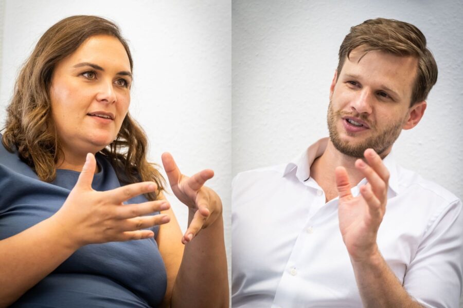 Zwei Menschen gestikulieren im Gespräch vor einem neutralen Hintergrund.