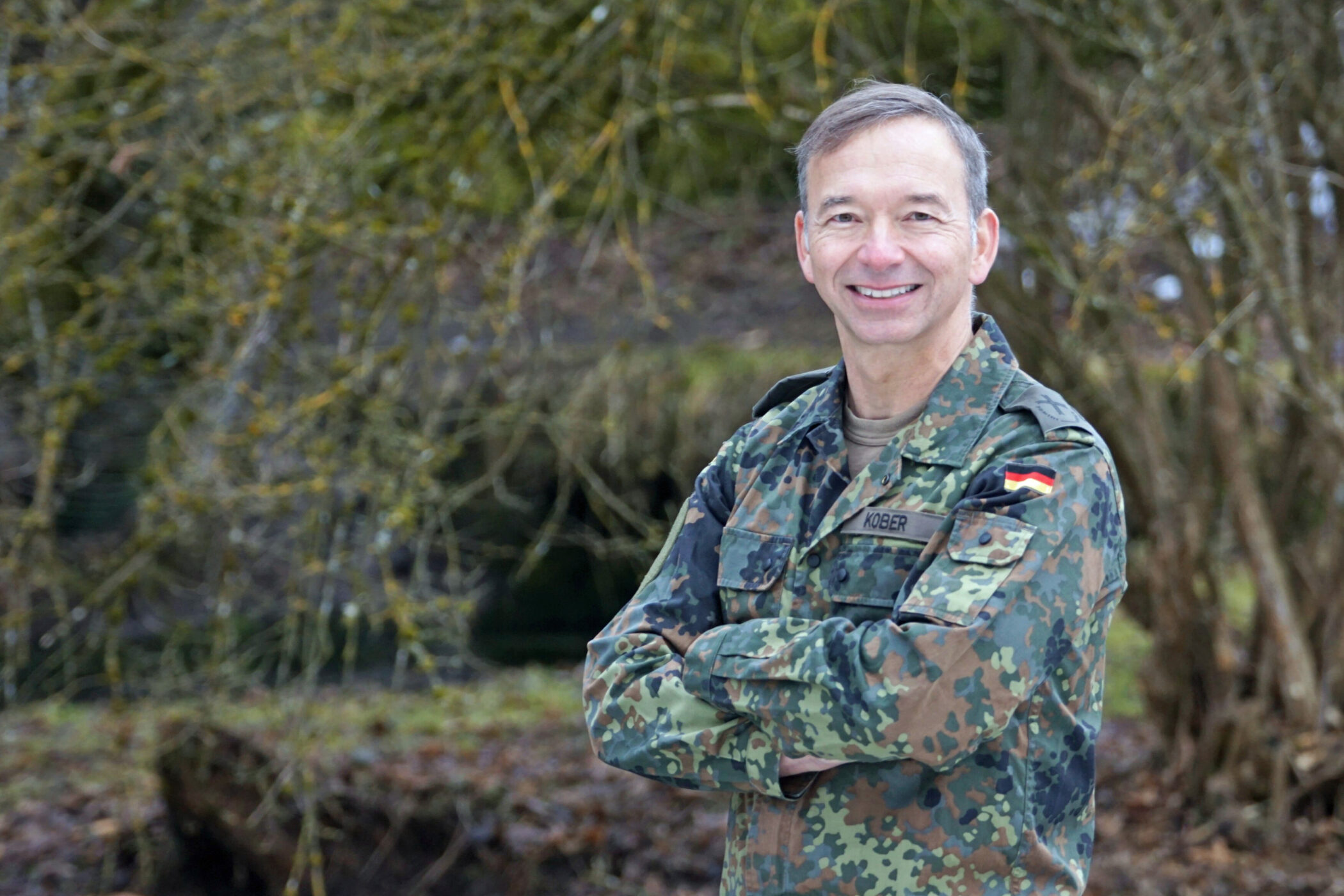 Person in Tarnuniform steht lächelnd im Freien, Arme verschränkt.