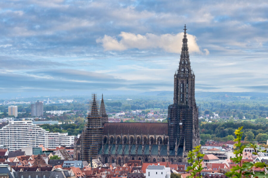 Große gotische Kirche mit Spitze, daneben Stadtansicht und bewölkter Himmel.