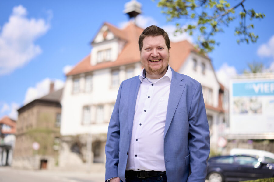 Mann im blauen Anzug vor unscharfem Haus und blauem Himmel.