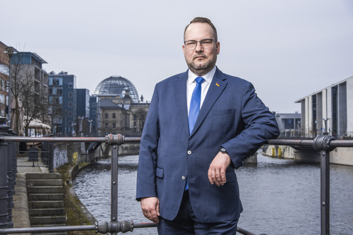 Ein Mann im Anzug steht an einem Geländer, im Hintergrund ein Fluss.