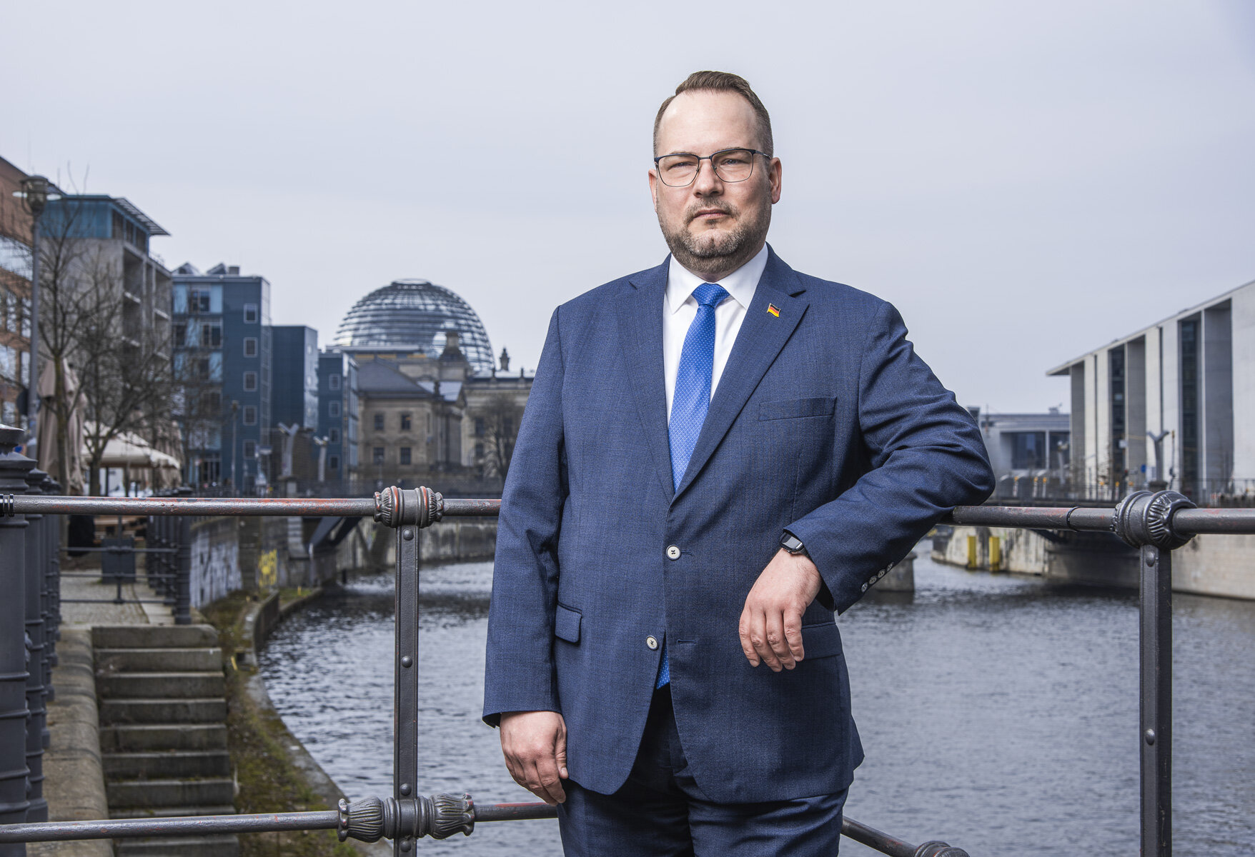 Ein Mann im Anzug steht an einem Geländer, im Hintergrund ein Fluss.