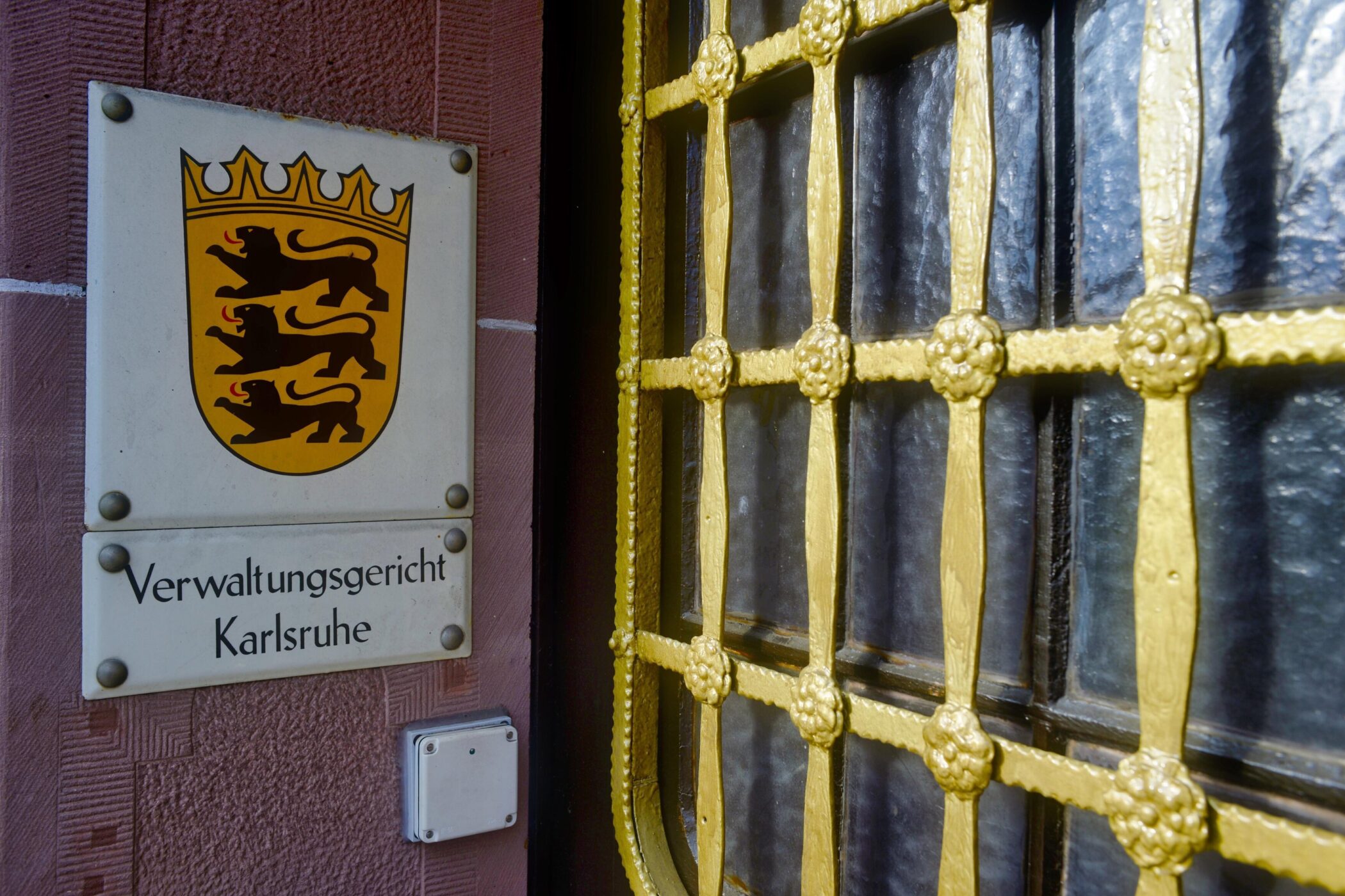 Schild mit Wappen und Text "Verwaltungsgericht Karlsruhe" neben vergittertem Fenster.