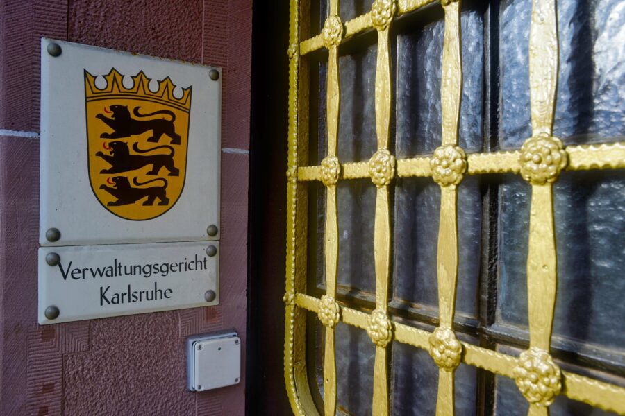 Schild mit Wappen und Text "Verwaltungsgericht Karlsruhe" neben vergittertem Fenster.