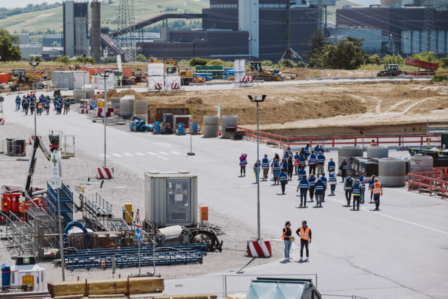 Gruppe mit Schutzwesten auf einer Baustelle, Industriegebäude im Hintergrund.