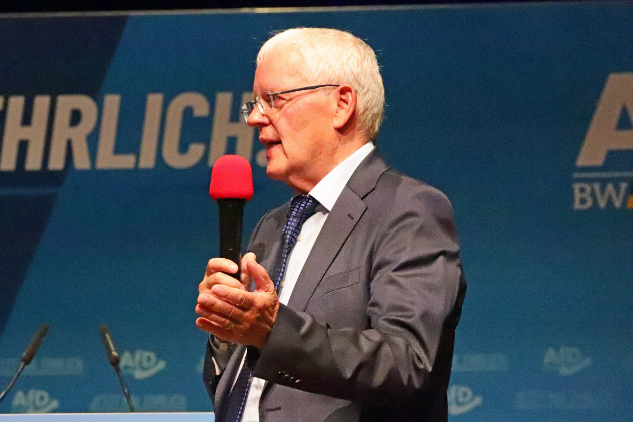 Älterer Mann im Anzug spricht in Mikrofon auf einer Bühne mit blauem Hintergrund.