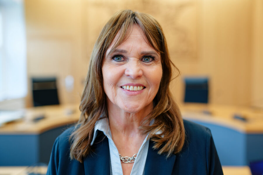 Frau mit langer brauner Haaren lächelt in einem Büro mit Schreibtischen.