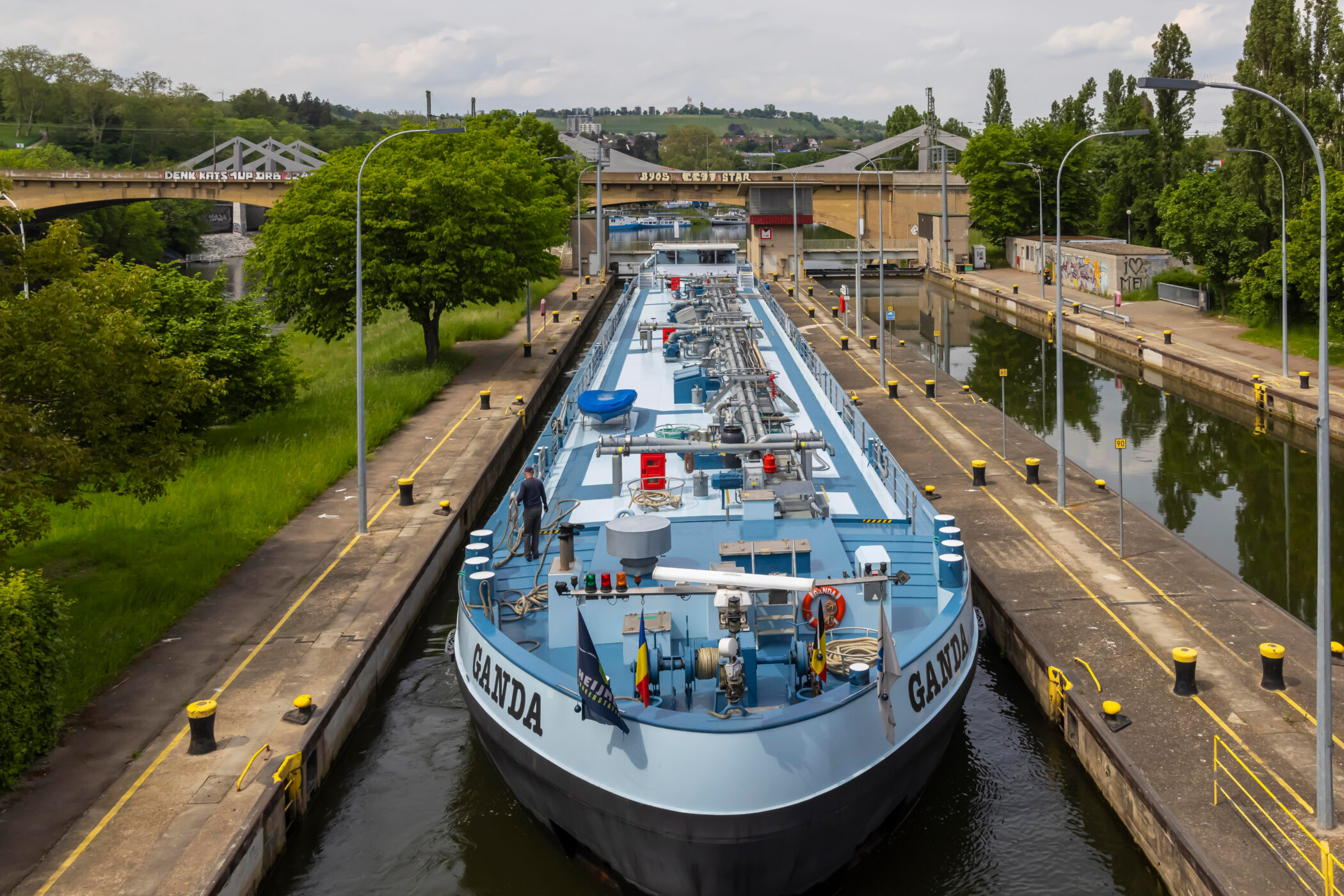 Großes Frachtschiff in Schleuse, Bäume, Brücke im Hintergrund. Schiffsschrift „GANDA“.