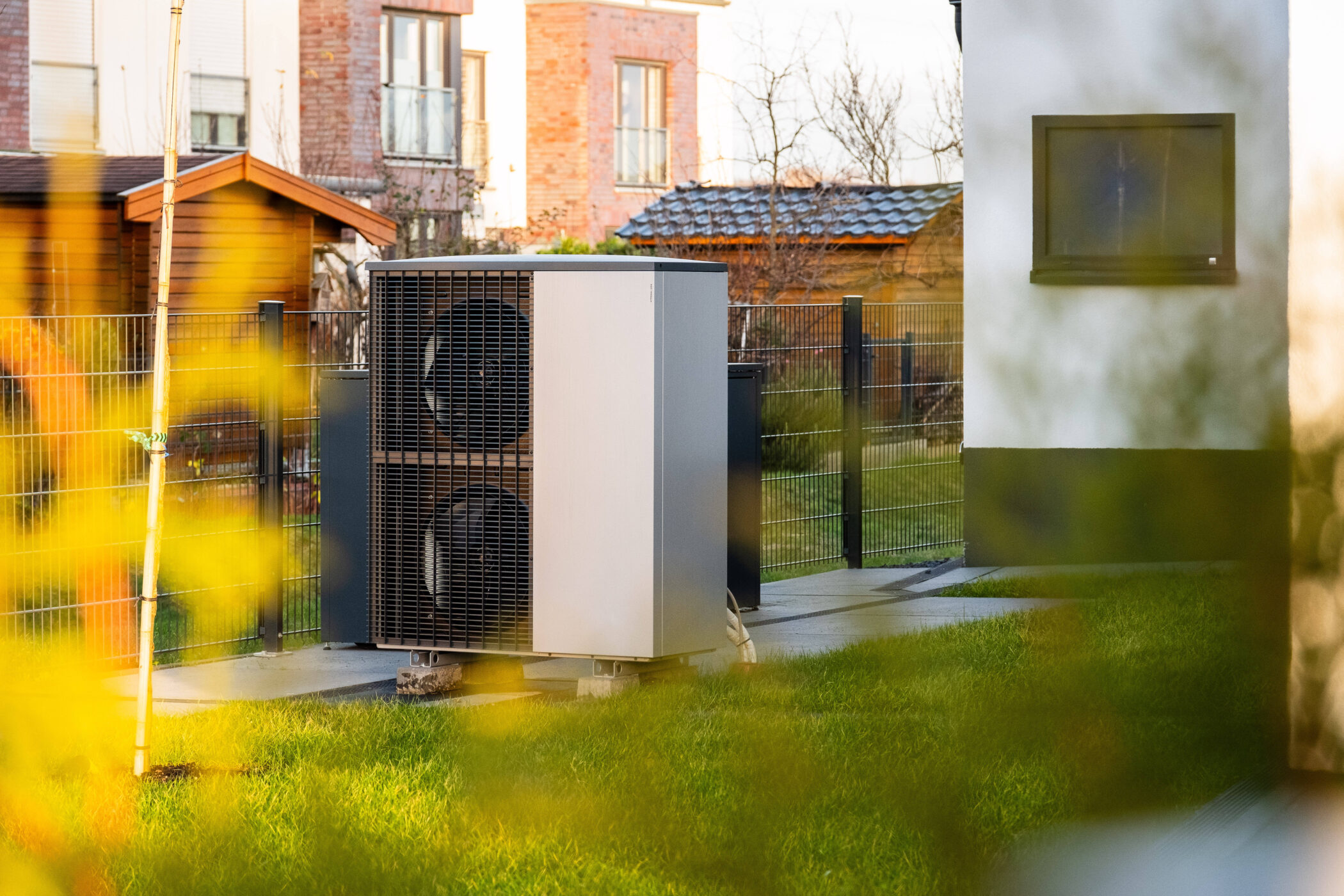 Wärmepumpe im Garten, umgeben von Rasen und Zaun, Häuser im Hintergrund.