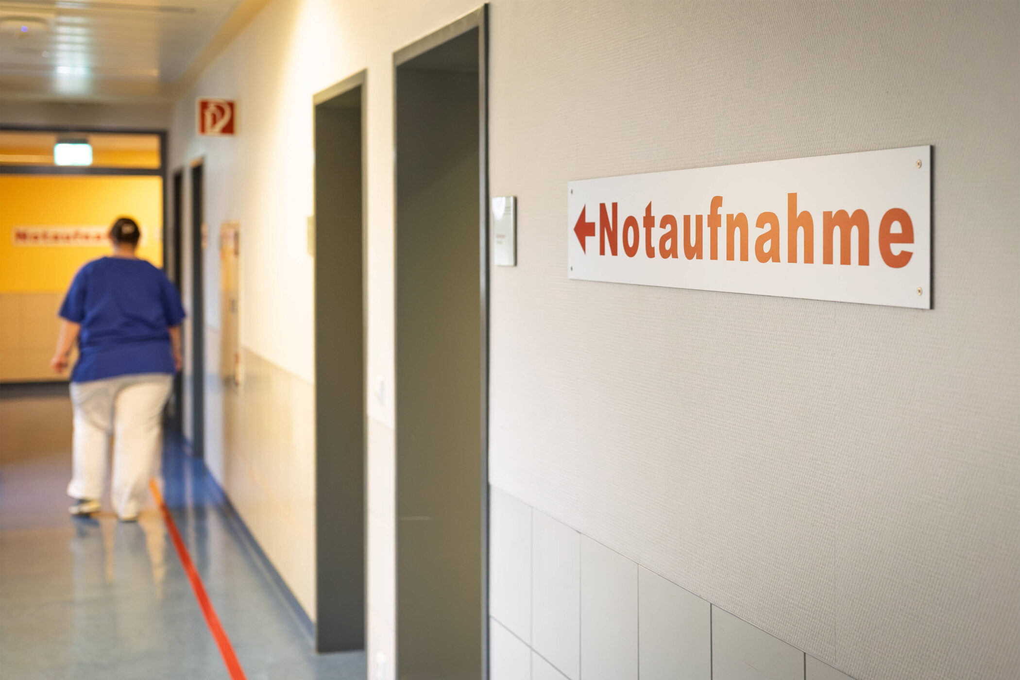 Flur mit Türschildern, rechts "Notaufnahme", eine Person geht im Hintergrund.