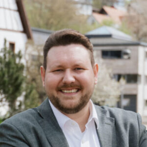 Lachender Mann im Anzug steht draußen, unscharfer Hintergrund mit Gebäuden und Bäumen.