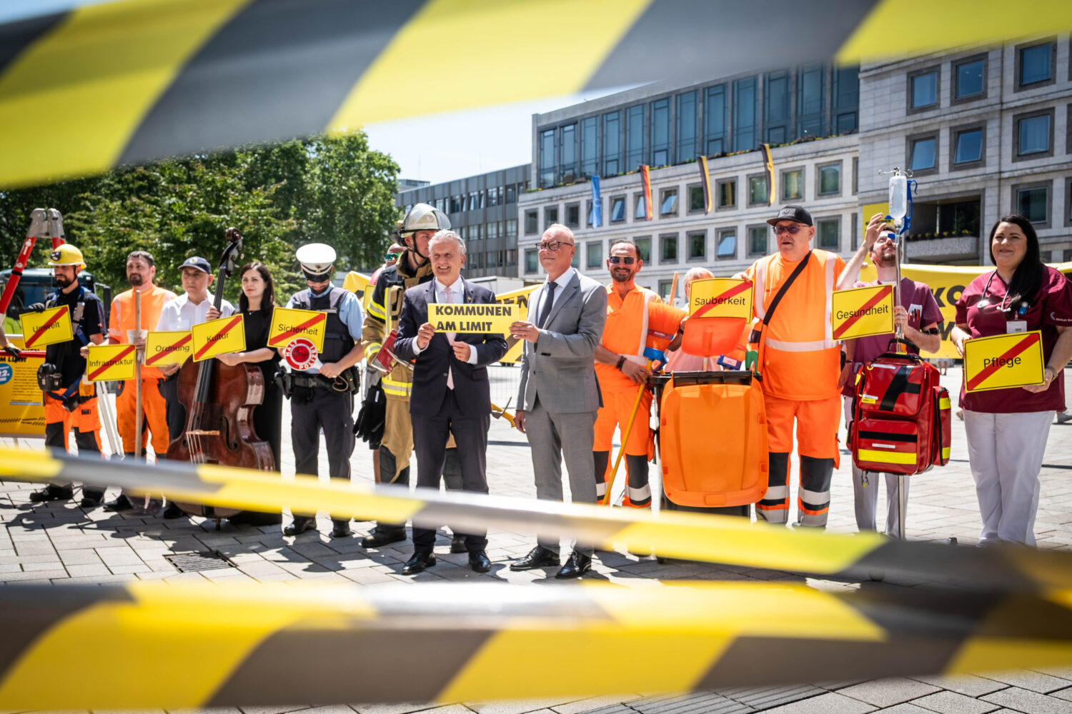 Gruppe von Menschen in Arbeitskleidung mit Schildern hinter Absperrband.