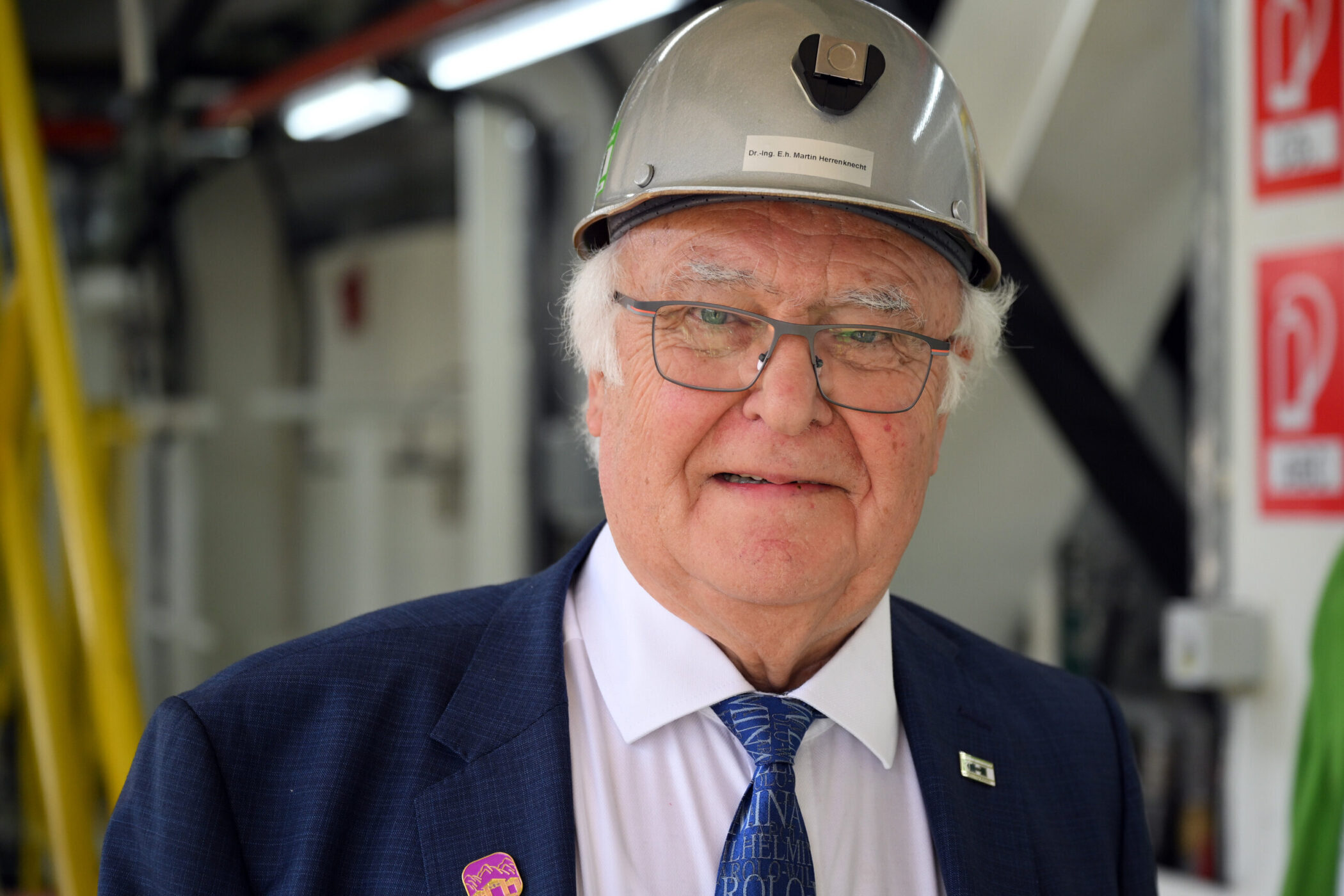 Älterer Mann mit Helm und Anzug in einem industriellen Umfeld.