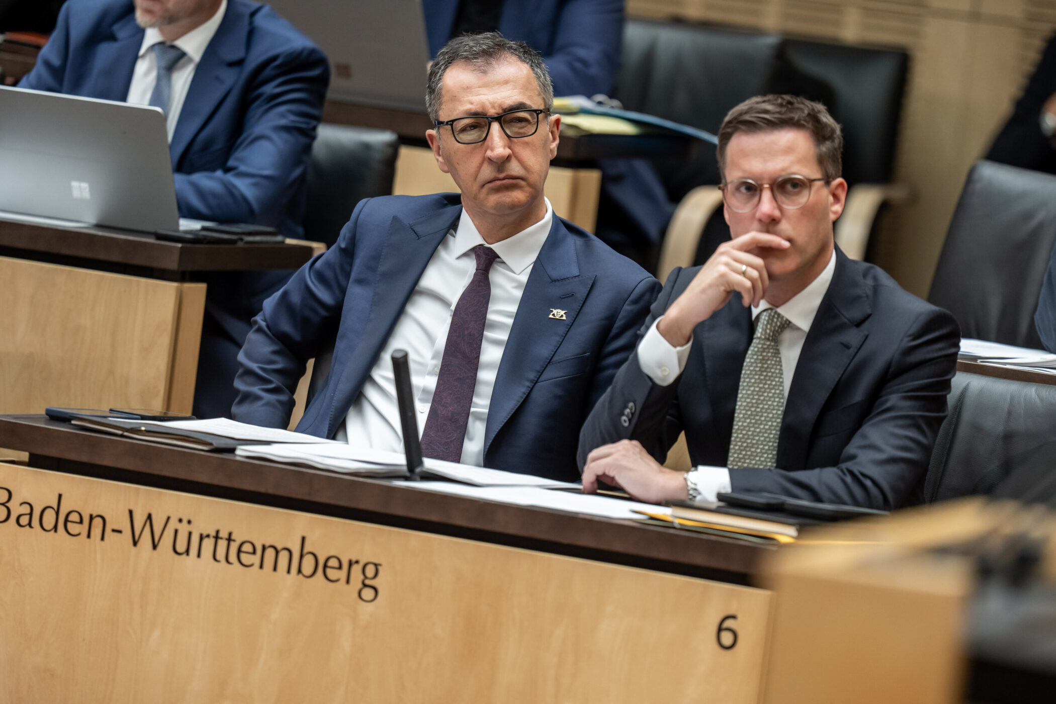 Zwei Männer in Anzügen sitzen, Schild "Baden-Württemberg 6" sichtbar.