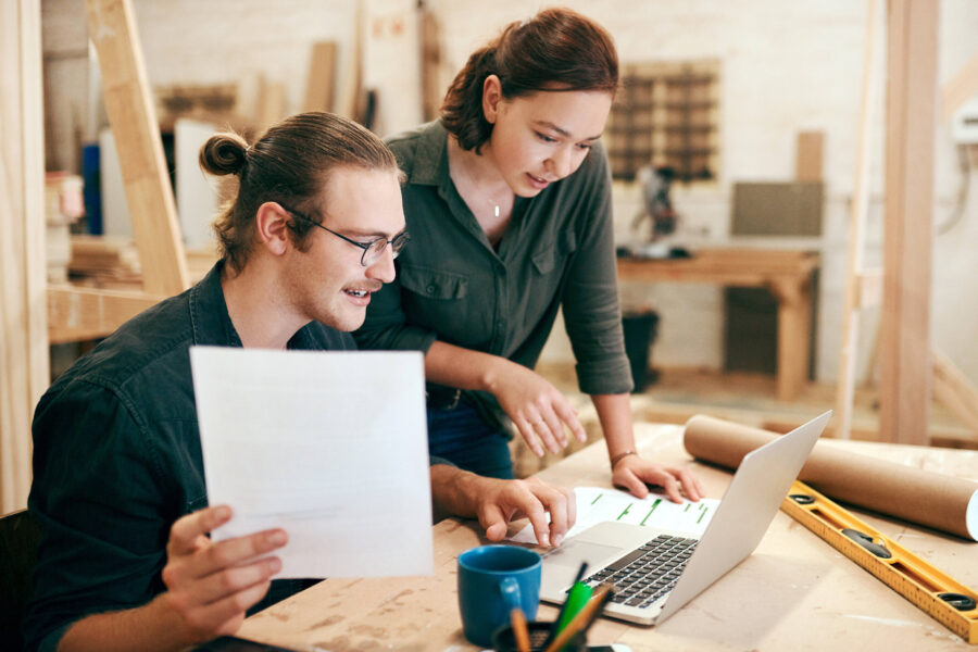 Zwei Personen betrachten ein Laptop, eine Person hält ein Papier. Tisch mit Werkzeug.