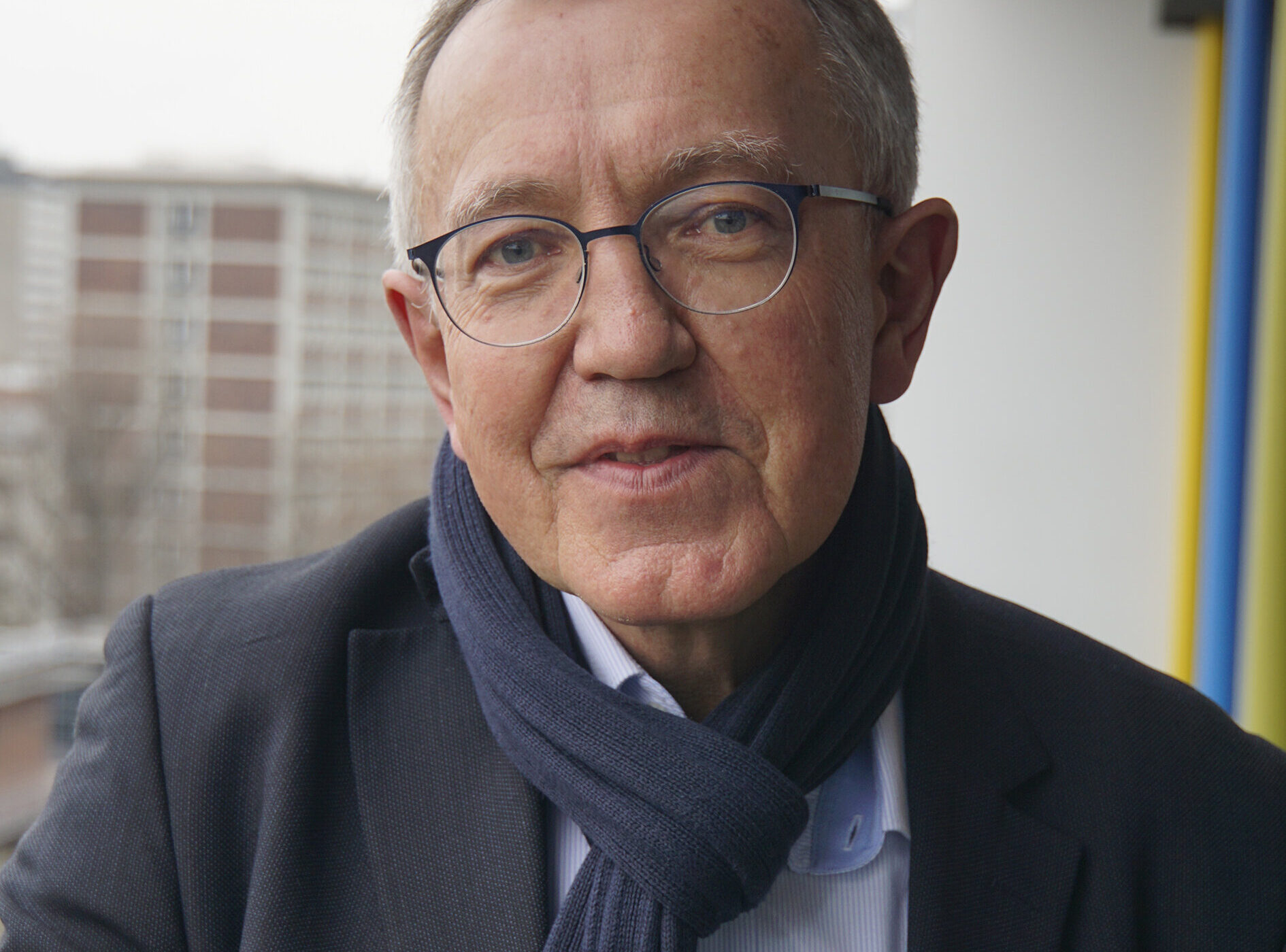 Älterer Mann mit Brille und Schal steht auf einem Balkon, Gebäude im Hintergrund.