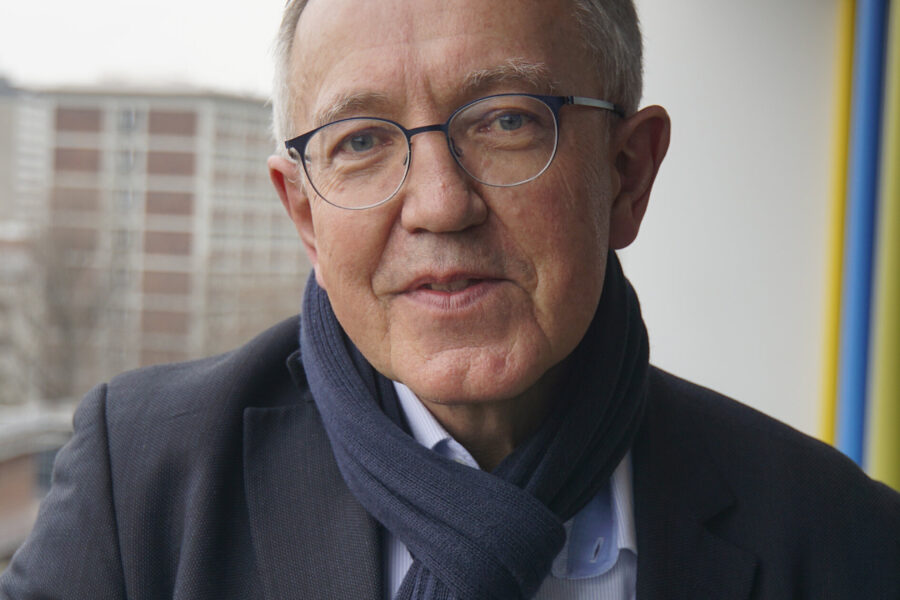 Älterer Mann mit Brille und Schal steht auf einem Balkon, Gebäude im Hintergrund.