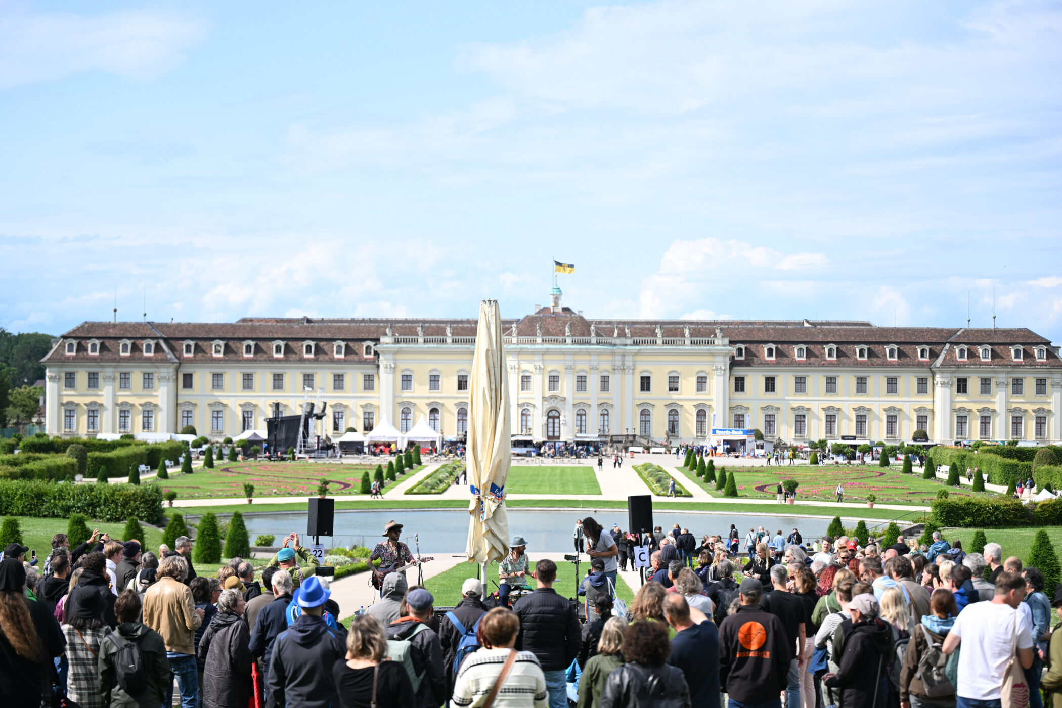 Menschen vor Schloss, Garten mit Wegen, Flagge auf Dach, sonniges Wetter.