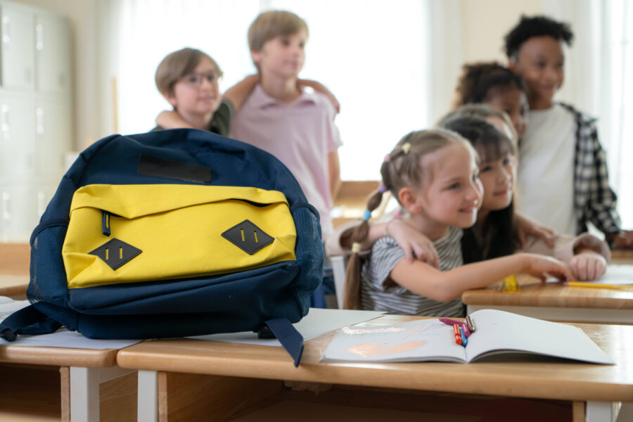 Blauer und gelber Rucksack auf Tisch, dahinter sieben Kinder in einem Klassenraum.