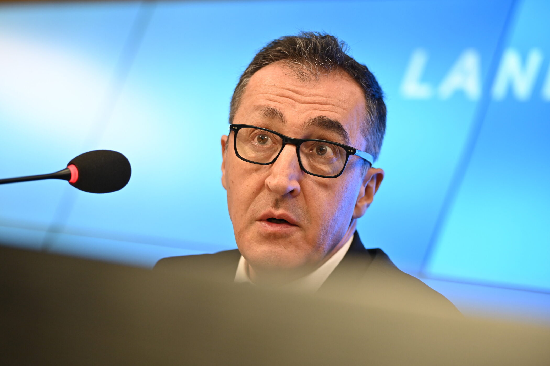 Ein Mann mit Brille spricht in ein Mikrofon vor blauem Hintergrund.