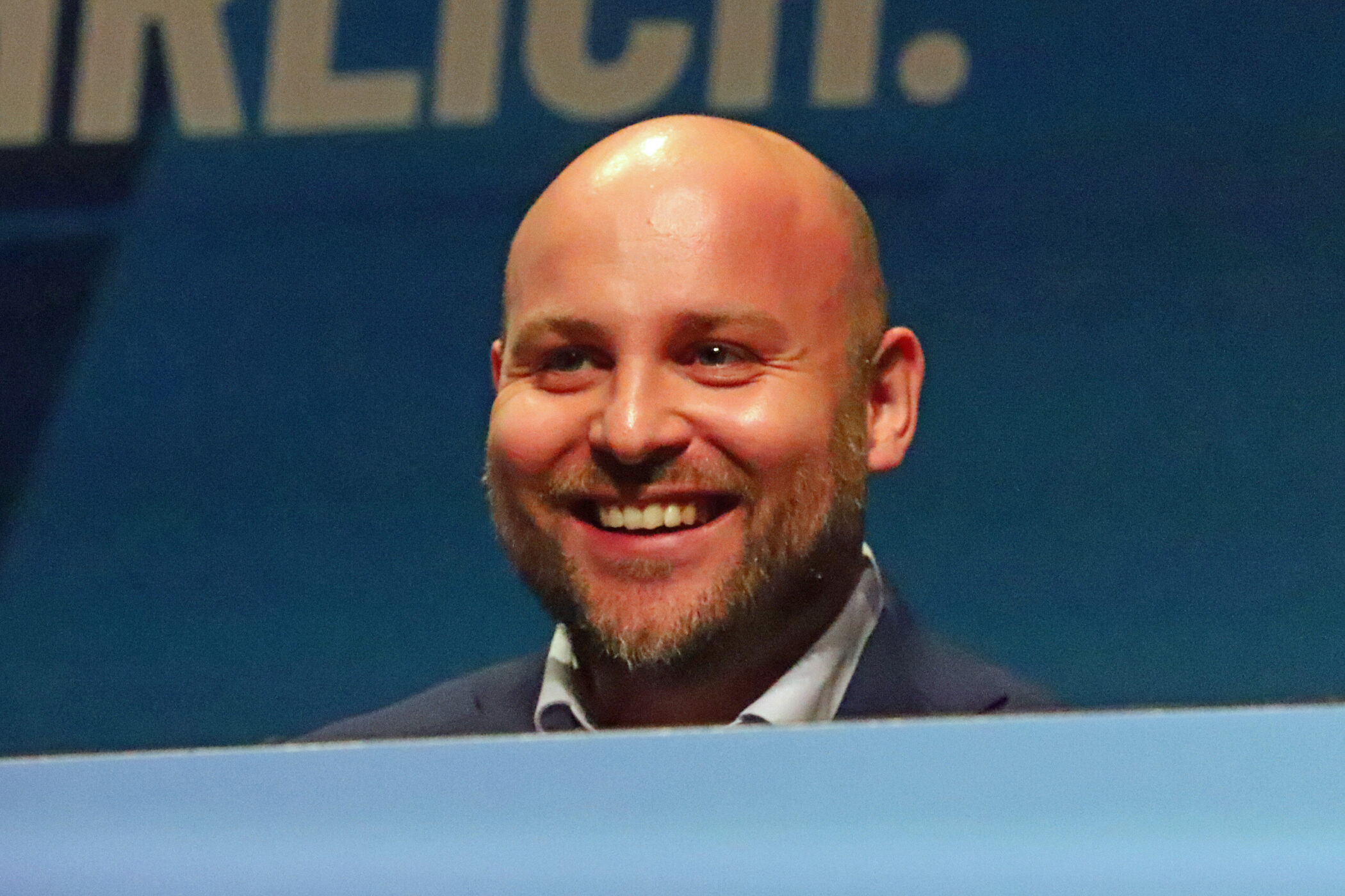 Lächelnder Mann mit Glatze im Anzug, Hintergrund mit unleserlichem Text.