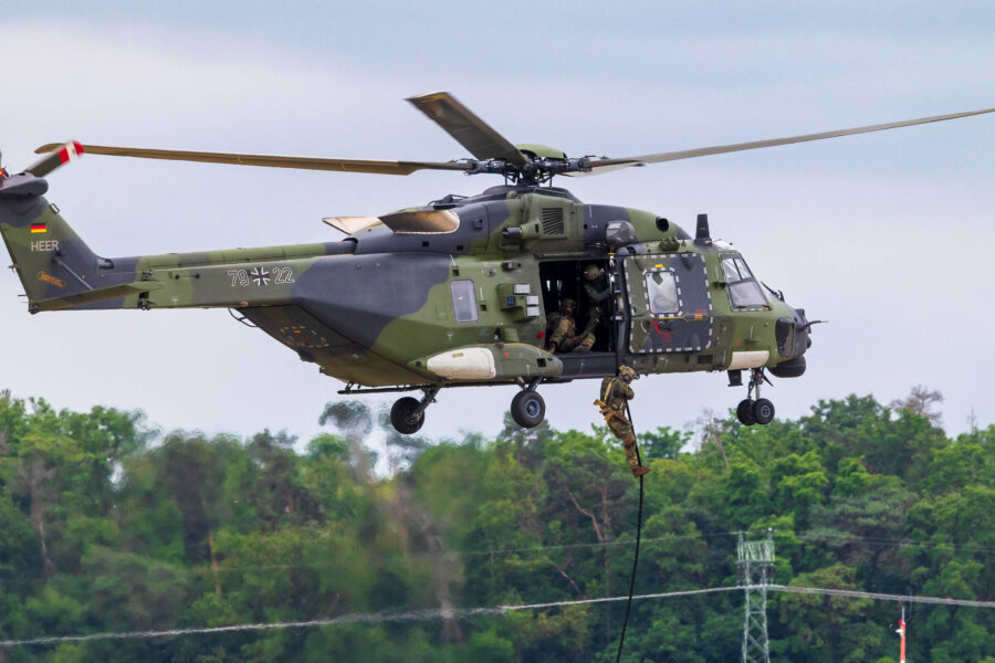 Grüner Militärhubschrauber, zwei Soldaten seilen sich ab, Wald im Hintergrund.